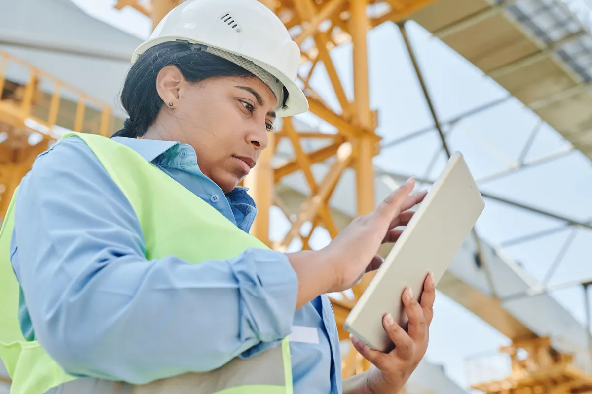 A worker in a hard hat and hi-vis vest using a tablet near a crane