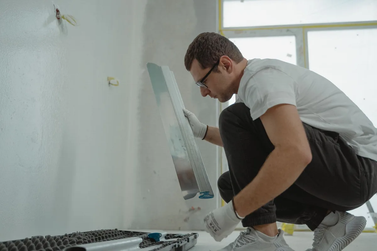 A man in a white T-shirt plastering an interior wall with a long trowel