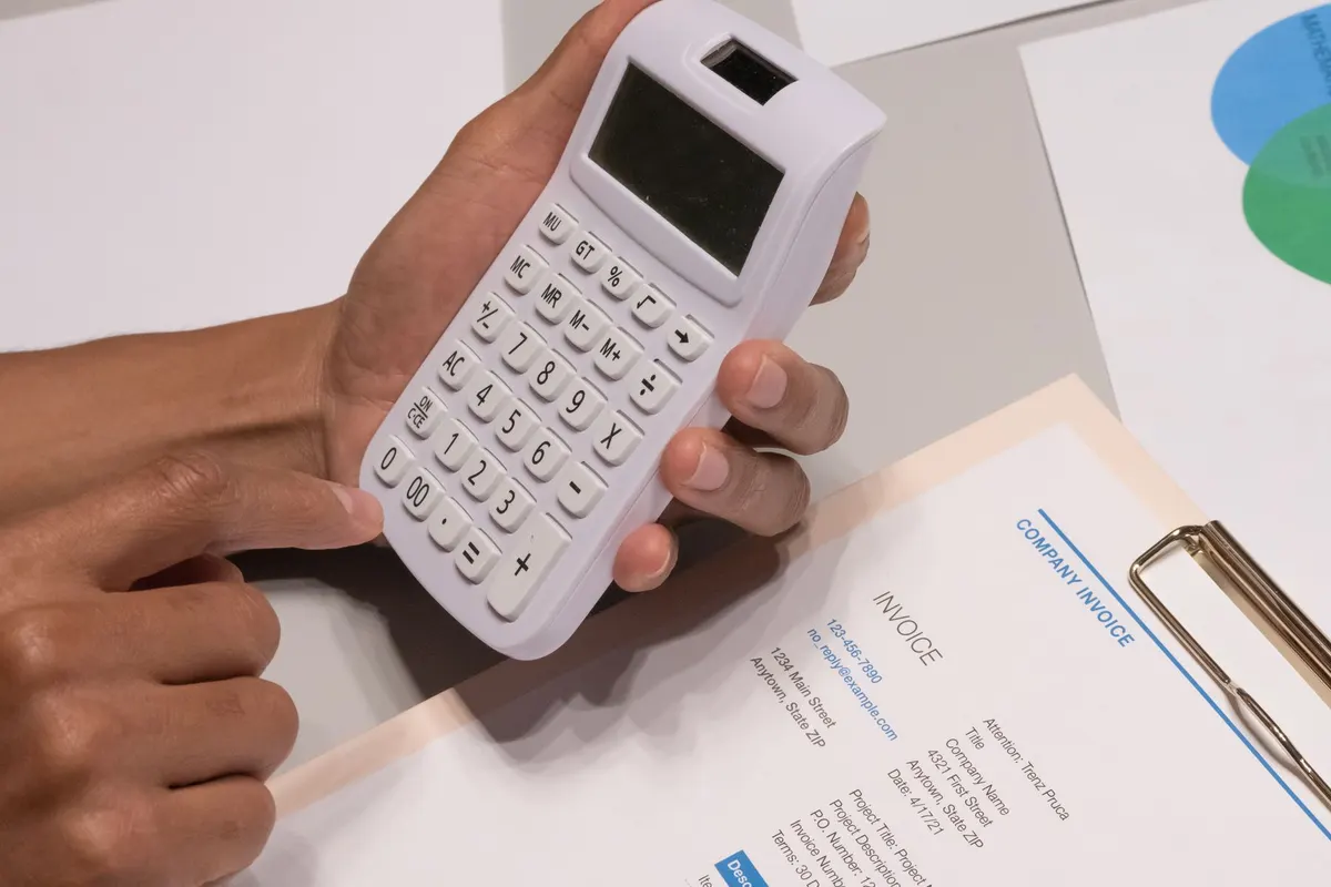 A hand holding a calculator over a printed invoice on a clipboard