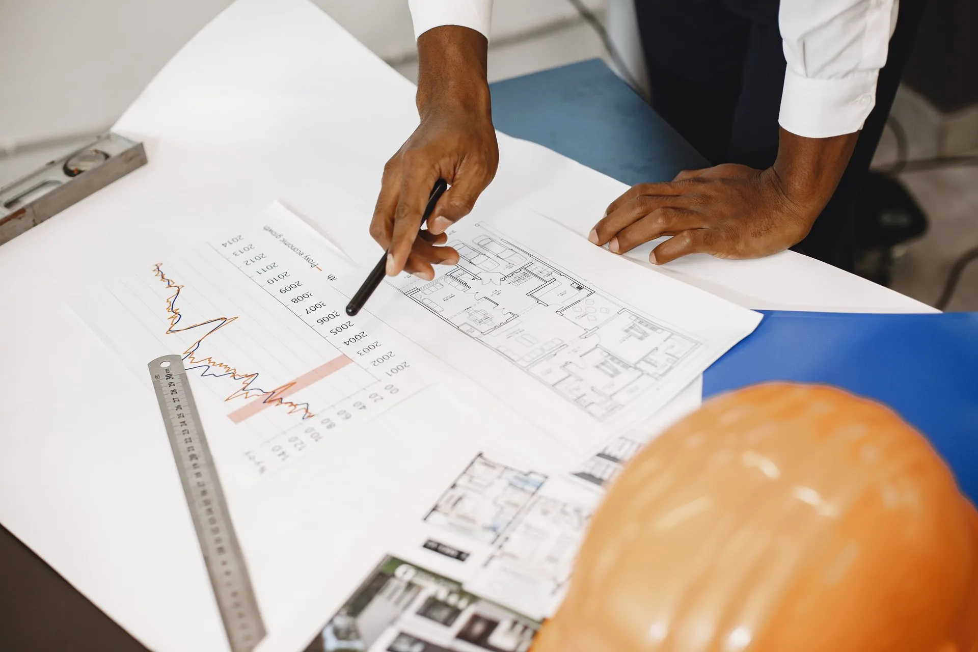 A builder's hand pointing at a price trend chart laid over residential floor plans, with a hard hat on the desk