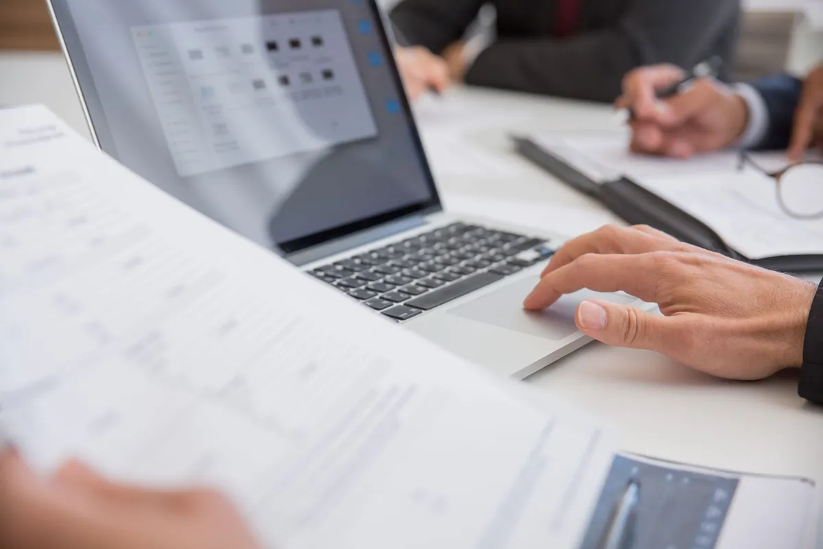 Hands working with printed documents beside a laptop on an office desk
